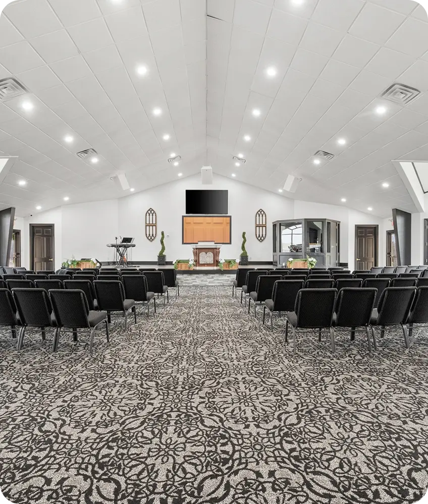 A modern sanctuary with rows of black chairs, a vaulted ceiling, and an ornate black-and-white patterned carpet leading to a stage.